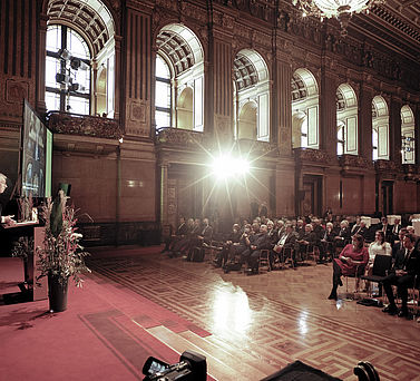 Eine Weitwinkel-Ansicht der Preisverleihung im großen Saal des Hamburger Rathaus. Akademiepräsident Prof. Dr. Edwin J. Kreuzer steht links hinter dem Rednerpult, während das Publikum rechts auf Stühlen sitzt.