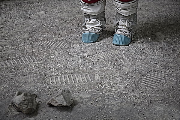 Ein sandiger Boden mit Schuhabdrücken und zwei kleinen Steinen im Vordergrund. Im Hintergrund sind Stiefel eines Raumanzugs zu erkennen.