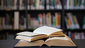 Zwei B&uuml;cher liegen ge&ouml;ffnet &uuml;bereinander. Unscharf im Hintergrund stehen gef&uuml;llte B&uuml;cherregale einer Bibliothek.