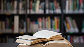 Zwei B&uuml;cher liegen ge&ouml;ffnet &uuml;bereinander. Unscharf im Hintergrund stehen gef&uuml;llte B&uuml;cherregale einer Bibliothek.