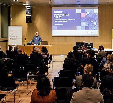 Prof. Dr. Alex van Belkum steht hinter dem Rednerpult und spricht zum Publikum. Hinter ihm auf einer Leinwand ist der Titel seines Vortrags &bdquo;Future Diagnostics Today&ldquo; zu lesen. Einige der Teilnehmenden heben ihre H&auml;nde, um Fragen oder Anmerkungen beizutragen.