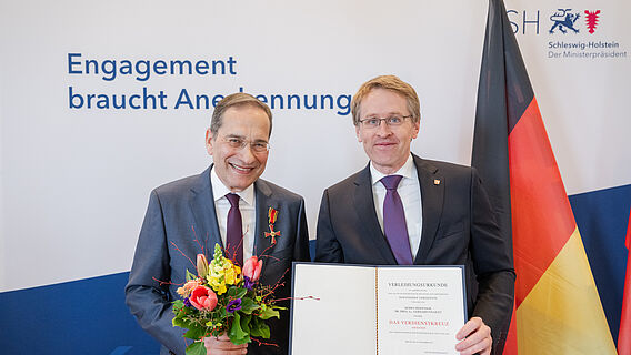 Akademiemitglied Gerhard Fouquet mit Bundesverdienstkreuz am Revers seiner Anzugjacke steht neben Schleswig-Holsteins Ministerpr&auml;sident Daniel G&uuml;nther. Rechts im Hintergrund ist die Flagge Deutschlands zu sehen.
