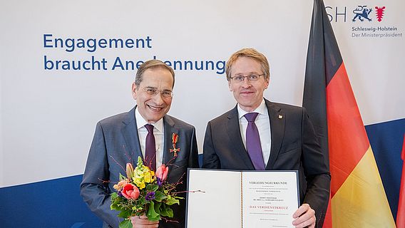Akademiemitglied Gerhard Fouquet mit Bundesverdienstkreuz am Revers seiner Anzugjacke steht neben Schleswig-Holsteins Ministerpr&auml;sident Daniel G&uuml;nther. Rechts im Hintergrund ist die Flagge Deutschlands zu sehen.