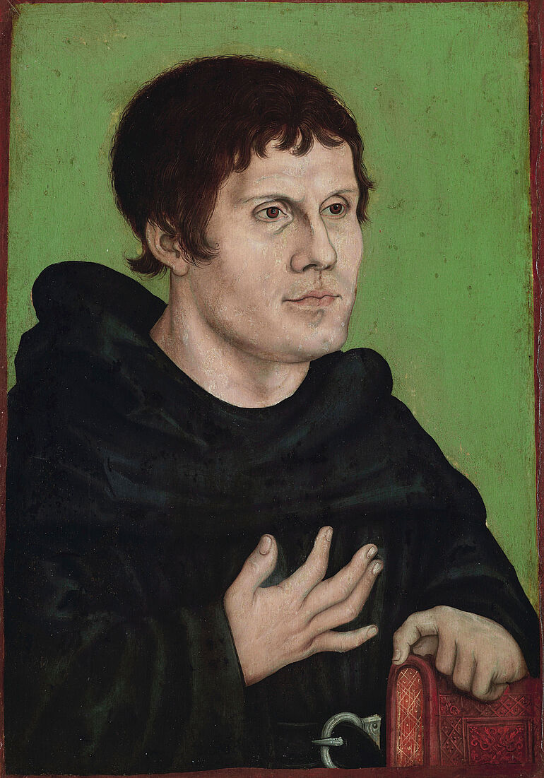 Das gemalte Portrait zeigt Martin Luther in einer M&ouml;nchskutte vor einem gr&uuml;nen Hintergrund. Seine rechte Hand liegt beteuernd auf seiner Brust und die linke Hand auf einem in Leder gebundenen Buch. 