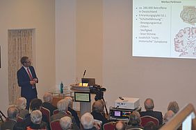 ein Gastsprecher tr&auml;gt vor einem Publikum in einem gro&szlig;en Saal einen Vortrag vor