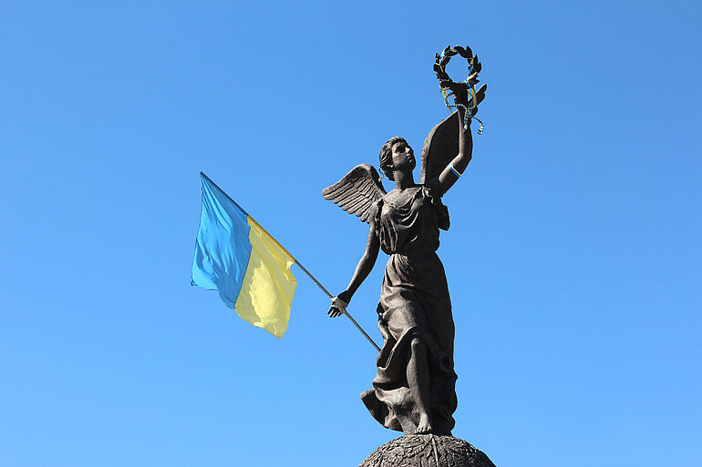 Die "Flying Ukraine"-Bronzestatue des Unabh&auml;ngigkeitsdenkmals vor einem blauen Himmel. Eine weibliche Gestalt verk&ouml;rpert mit ausgebreiteten Fl&uuml;geln die Unabh&auml;ngigkeit der Ukraine. In ihrer rechten Hand h&auml;lt sie die Flagge der Ukraine, w&auml;hrend sie mit der linken Hand einen Lorbeerkranz emporhebt.