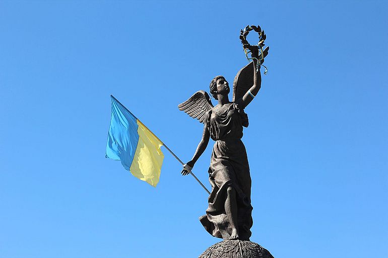 Die "Flying Ukraine"-Bronzestatue des Unabh&auml;ngigkeitsdenkmals vor einem blauen Himmel. Eine weibliche Gestalt verk&ouml;rpert mit ausgebreiteten Fl&uuml;geln die Unabh&auml;ngigkeit der Ukraine. In ihrer rechten Hand h&auml;lt sie die Flagge der Ukraine, w&auml;hrend sie mit der linken Hand einen Lorbeerkranz emporhebt.
