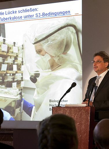 Preistr&auml;ger des Hamburger Wissenschaftspreises 2009 Prof. Dr. Stefan Ehlers steht hinter dem Rednerpult. Auf einer Leinwand hinter ihm ist eine Person im Schutzanzug zu sehen, die einen Klarsichtbeh&auml;lter mit experimentellen Tuberkulosekulturen in den H&auml;nden h&auml;lt.