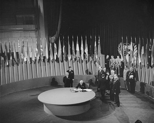 Ein schwarz-weißes Bild zeigt den Moment der Unterzeichnung der Charta der Vereinten Nationen im Rahmen der Konferenz von San Francisco (26. Juni 1945). Die Vertreter der Mitgliedsländer stehen in einem runden Raum, der von den Flaggen der Mitgliedsländer umgeben ist, während der Repräsentant der USA die Charta an einem großen runden Tisch unterzeichnet.