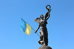 Die "Flying Ukraine"-Bronzestatue des Unabhängigkeitsdenkmals vor einem blauen Himmel. Eine weibliche Gestalt verkörpert mit ausgebreiteten Flügeln die Unabhängigkeit der Ukraine. In ihrer rechten Hand hält sie die Flagge der Ukraine, während sie mit der linken Hand einen Lorbeerkranz emporhebt.