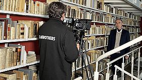Projektleiter Prof. Dr. Christian Brockmann steht neben einem B&uuml;cherregal vor der Kamera bei den Dreharbeiten in der Herzog August Bibliothek in Wolfenb&uuml;ttel. 