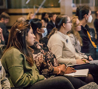 Eine Nahaufnahme vom Publikum in der ersten Reihe. Zwei der sechs zu sehenden Teilnehmer: innen tragen einen Mund-Nasen-Schutz.