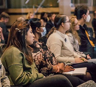 Eine Nahaufnahme vom Publikum in der ersten Reihe. Zwei der sechs zu sehenden Teilnehmer: innen tragen einen Mund-Nasen-Schutz.