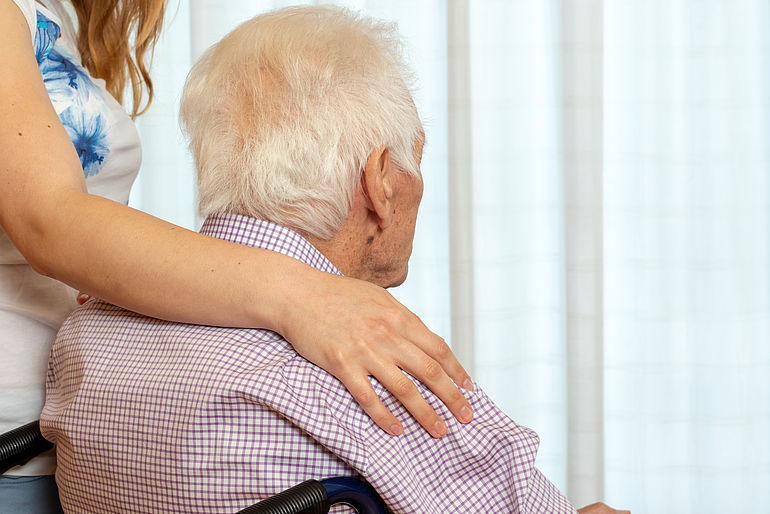 Der Arm einer Frau liegt auf der Schulter eines im Rollstuhl sitzenden älteren Mannes, der dem Betrachter den Rücken zukehrt.