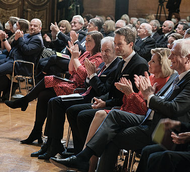Der Preisträger Prof. Dr. Dr. Fabian Theis inmitten von applaudierendem Publikum. Er sitzt in der vordersten Reihe neben Wissenschaftssenatorin Katharina Fegebank, Prof. Dr. Edwin J. Kreuzer und Eva-Maria Greve.