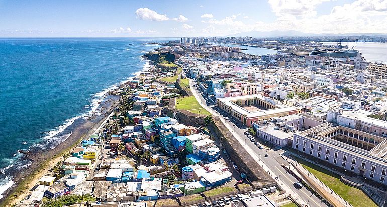 Eine Aufnahme aus der Vogelperspektive. Links liegt der Slum La Perla mit improvisierten H&uuml;tten direkt am Meer. Der Slum ist durch eine breite Mauer von den rechts liegenden gepflegten und teils luxuri&ouml;s wirkenden Geb&auml;uden getrennt.